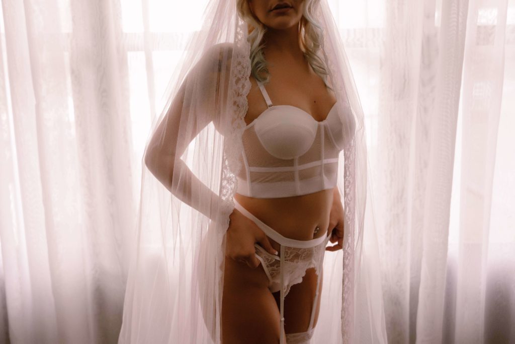 bride in lingerie and garters, wearing a long veil and standing in front of a curtained window during a bridal boudoir shoot