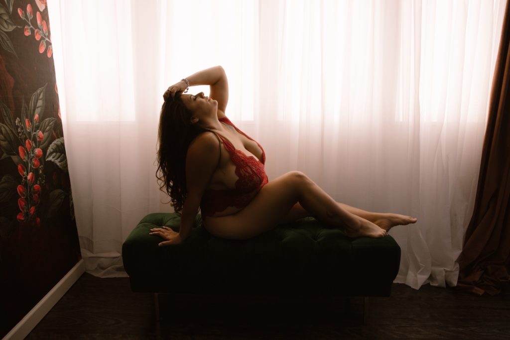 woman wearing red lingerie reclining on a green bench in front of a white curtained window