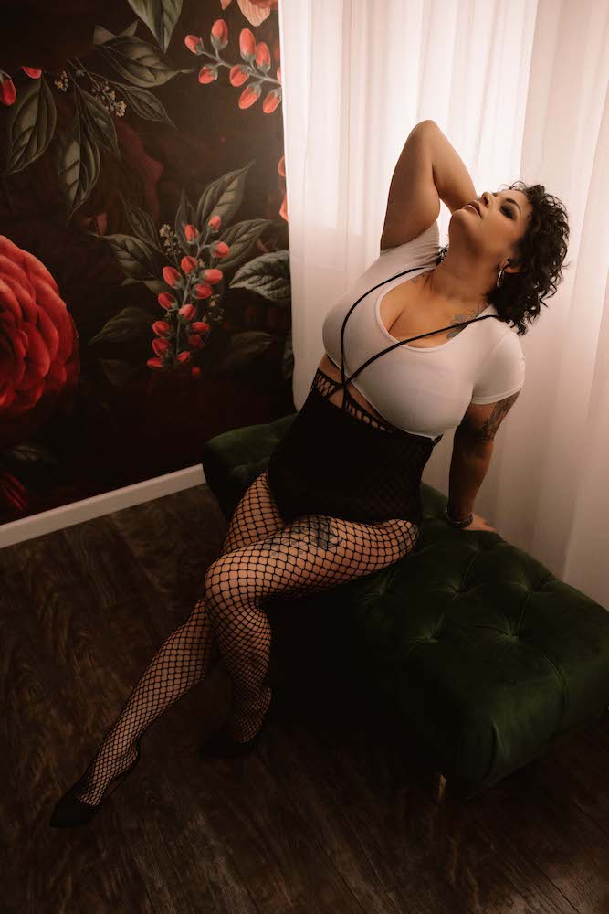 Woman in fishnet stockings and a white crop top poses in on a green bench in front of a window and a floral-covered wall